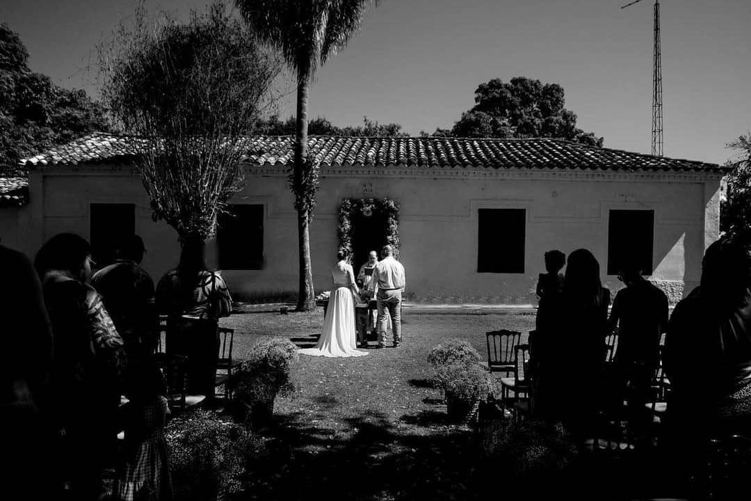 Fotografia em preto e branco de uma cerimônia diante da casa, com os convidados à sombra