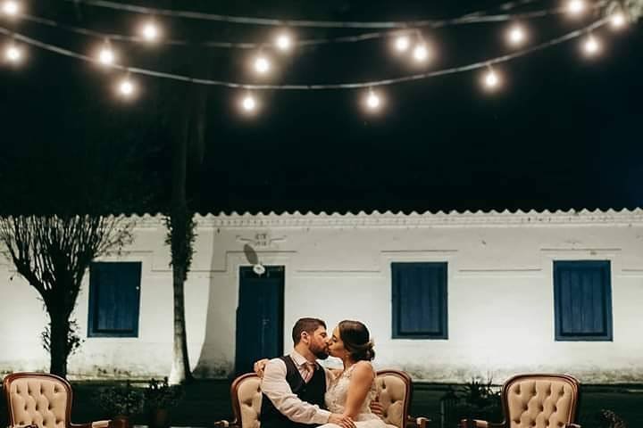 Os noivos sob o varal de lâmpadas, diante da fachada iluminada à noite