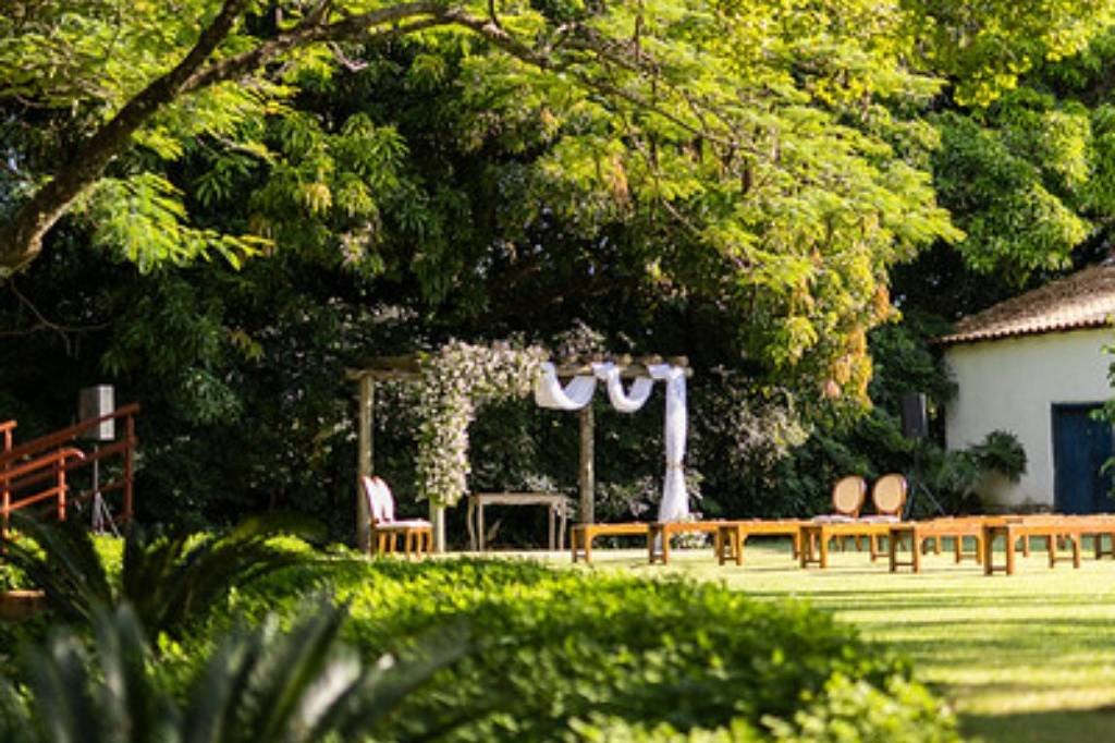 Altar com cortinas brancas sob as árvores, bancos no gramado e a casa à direita